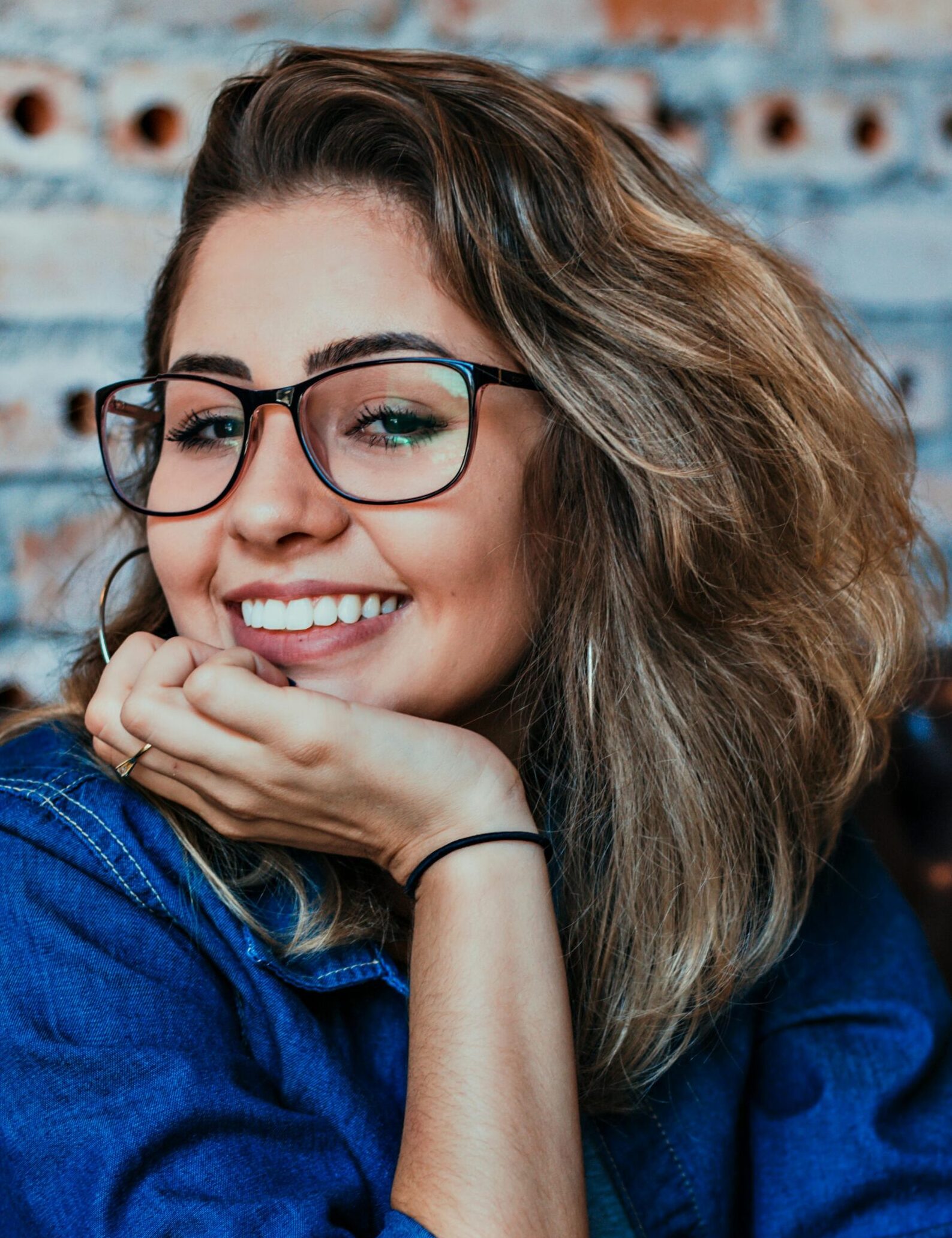 A young woman wearing glasses and denim, smiling in an indoor setting.